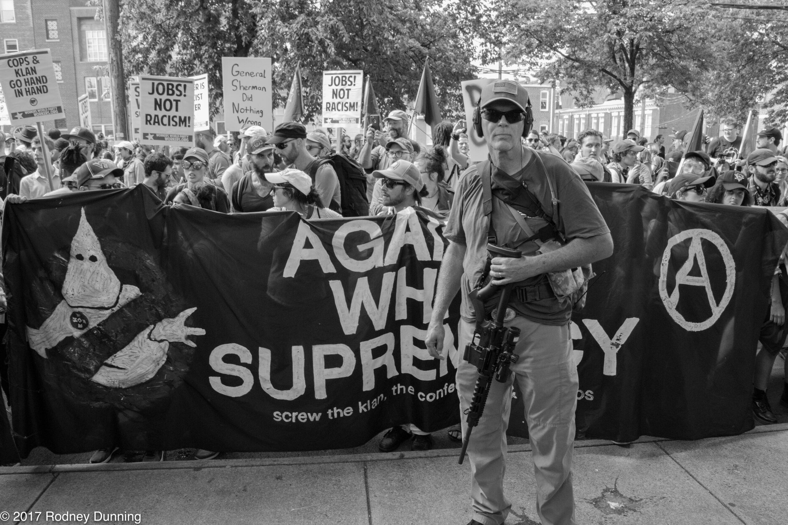 The image depicts a protest with a large group of people holding signs and banners. The main banner in the foreground reads "AGAINST WHITE SUPREMACY screw the klan, the confederacy, and cops" with an illustration of a Ku Klux Klan member crossed out. Another prominent sign reads "JOBS! NOT RACISM!" There are several other signs visible in the background" A person in front of the banner is holding what appears to be a firearm. The image is in black and white and has been credited to Rodney Dunning (2017).