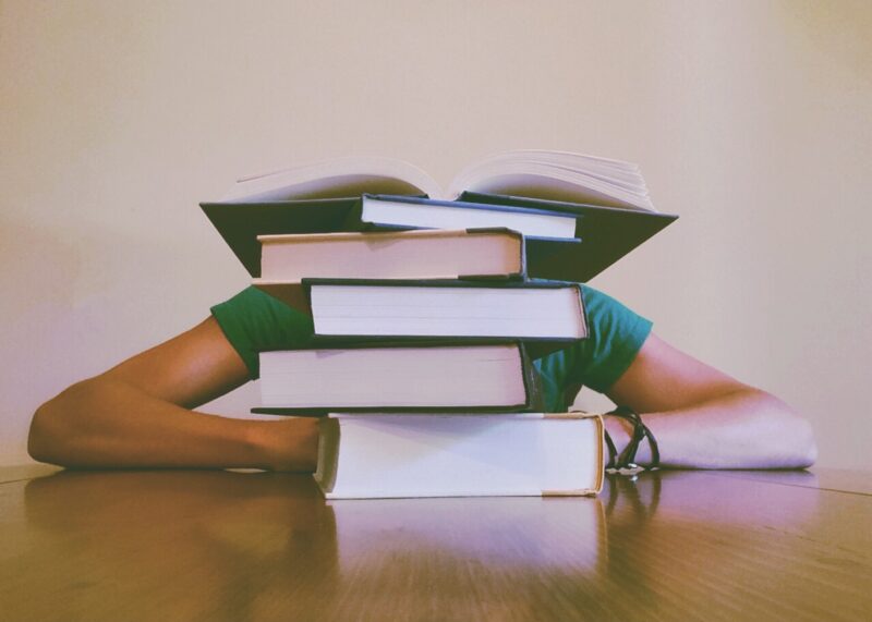 Image of someone leaning against a table with their head down, obscured by a pile of four closed books, with an open book at the top