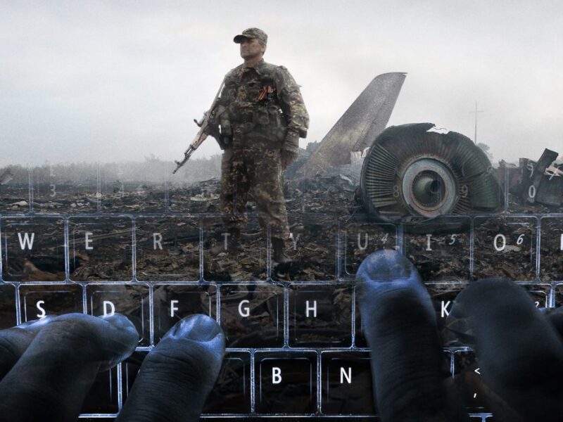 The image shows a soldier standing in a wreckage site with debris and parts of an aircraft visible in the background. The foreground features hands typing on a keyboard, creating an overlay effect with the scene behind it.