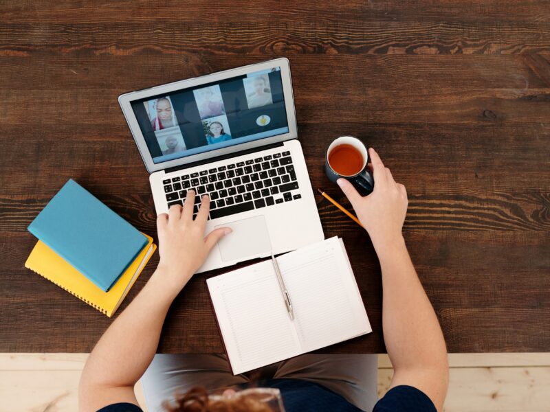 The image shows a person sitting at a wooden table, using a laptop for a video call. The laptop screen displays four participants in the video call. The person has an open notebook with a pen placed diagonally on it, and is holding a cup of tea or coffee in the other hand. There are also two closed notebooks, one blue and one yellow, placed on the table next to the laptop.