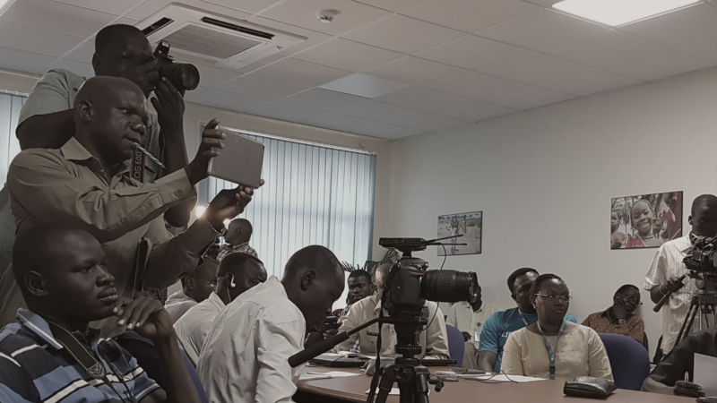 The image shows a group of people in a room, some of whom are taking photographs or recording videos. There are several cameras and recording devices visible.The room has fluorescent lighting and there is a window with vertical blinds in the background. There are also some posters on the walls.