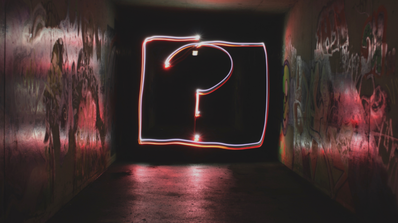 The image shows a dark tunnel with graffiti on the walls. In the center of the tunnel, there's a large question mark outlined with light, creating a glowing effect. The question mark is enclosed within a rectangular frame also made of light.