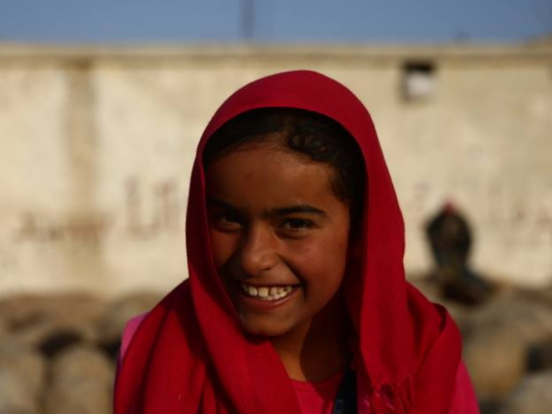 The image shows a person wearing a red headscarf and a pink shirt standing in front of a wall with some text on it.