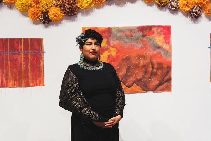 The image shows a person standing in front of a wall decorated with colorful paper flowers and two pieces of artwork. The person is wearing a black outfit with intricate patterns and a beaded necklace.